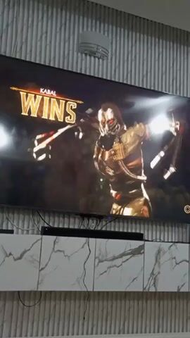Here's a description of the video:

A TV screen displays the end of a Mortal Kombat match. The character Kabal is declared the winner. The screen then transitions to a close-up of Kabal, with special effects. The TV is mounted on a white and gray cabinet with a marble-like pattern. Above the TV is a wall with gray vertical stripes and a small round light fixture. The room appears to be indoors.