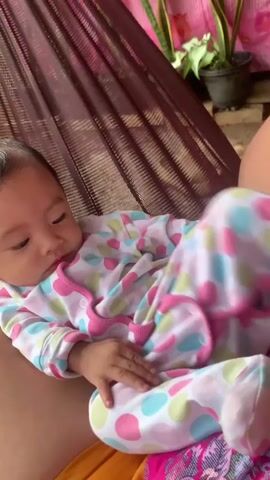 A baby in a polka-dotted onesie is lying in a hammock. The baby's eyes are closed and then open. The baby then begins to cry and moves their arms. The baby's expression changes from crying to a more neutral face. The background shows a plant and part of a hammock.