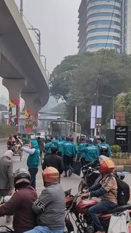 The video shows a busy street scene in what appears to be a city, likely in Bangladesh, based on the language on a bus and the apparel of some pedestrians. The scene unfolds under an elevated metro track. A group of individuals wearing similar teal jackets with the word "police" on them are walking across the street, potentially in a parade or demonstration. Several people on motorcycles are waiting at the intersection, along with a bicycle rickshaw and other vehicles. A large bus is visible in the background, along with several high-rise buildings, giving an impression of a bustling urban environment. The sky is overcast, indicating a day with moderate light.