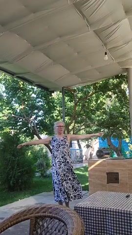 A middle-aged woman with blonde hair, wearing glasses and a long black and white patterned dress, stands on a patio, arms extended wide. She is outdoors, under a white canopy supported by metal poles. Behind her, trees and greenery are visible. A wicker chair is in the foreground, and a table with a checkered cloth is to the right. The woman appears to be dancing or posing.