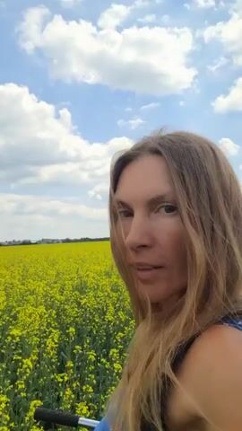 A selfie-style video shows a woman with long blonde hair in a field of yellow flowers under a blue sky with white clouds. The woman is looking at the camera and smiling. The field appears to be a canola field.