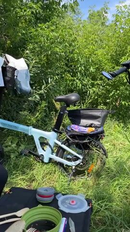The video shows a scenic outdoor setting with two electric bicycles parked amidst lush green foliage, likely a park or a nature trail. The bicycles are equipped with bags and baskets, suggesting a picnic or a camping trip. In the foreground, a picnic mat is laid out with food containers and cutlery, indicating that a meal is in progress or about to begin. The background features a calm river or lake, surrounded by trees, under a clear blue sky with scattered clouds. The overall atmosphere is peaceful and serene, perfect for an outdoor adventure.