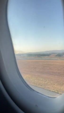 The video shows a view from an airplane window looking out at an airport tarmac during what appears to be takeoff or landing. The ground is a mix of dry, golden grass and paved surfaces with white lines. In the distance, there are buildings and hills under a clear blue sky. A thin plume of smoke or dust is visible on the horizon. The airplane window is oval-shaped and shows some smudges. The camera is positioned slightly to the left, looking out and forward. The lighting suggests it is either early morning or late afternoon.