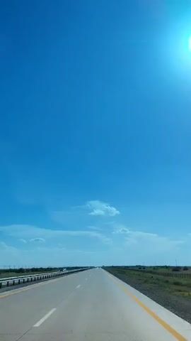 The video shows a view from a vehicle driving on a highway under a clear blue sky. The sun is visible in the upper right corner, casting a bright glare. The highway stretches out ahead, with a white line dividing the lanes and a yellow line along the right edge. To the left of the highway, there is a barrier and some faint objects that appear to be other vehicles in the distance. The right side of the highway is bordered by grass and some sparse vegetation. There are a few small clouds in the sky. The overall impression is of a long, open road on a sunny day.