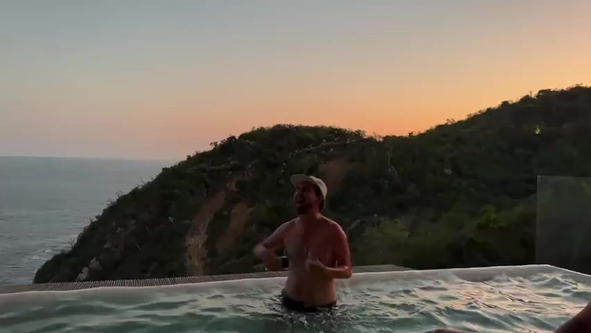 A man stands in a pool at sunset, holding a cell phone in his right hand. He wears a tan baseball cap, a necklace, and dark swim trunks. A green, hilly landscape is behind him, and the ocean is in the background. He appears to be talking and gesturing while holding the phone up like he's taking a selfie. The sky is a mix of orange, pink, and blue as the sun sets. The pool looks like an infinity pool, with a glass barrier on the edge.