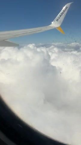 The video shows a view from the window of a Ryanair airplane, high above a thick layer of clouds. The sky is blue above the clouds. A portion of the plane's wing, bearing the Ryanair logo and a yellow accent, is visible. The window itself appears to be covered with a multitude of small droplets. The camera remains static throughout the brief clip.