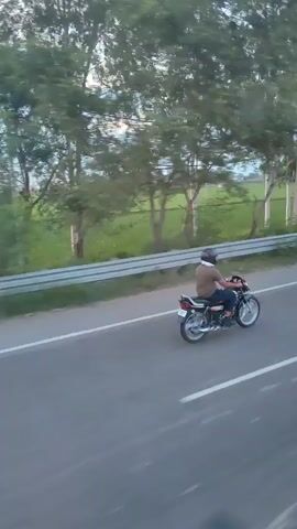 A man rides a motorcycle on a paved road. He is wearing a helmet and a t-shirt. The motorcycle is dark colored with chrome accents. The road is bordered by a white line on the right and a silver railing on the left. Beyond the railing, there is a grassy area with trees and fields. The sky is overcast with some sunlight breaking through. The camera appears to be following the motorcycle from the side.