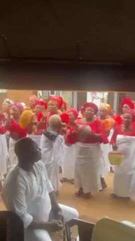The video shows a group of women in matching traditional attire performing a song. Most of them are wearing red tops, white wraps, and colorful headscarves, and they are holding musical instruments. The instruments appear to be shakers. They are standing in a loose group outside, under some sort of canopy. In front of them sits a man in a white outfit, watching the performance. Other people are visible in the background.