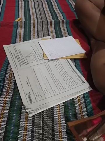 The video shows a person's hands interacting with a mobile phone and papers on a striped mat. The mat has red, white, green, and dark blue stripes. In the foreground, several papers are stacked. The top paper is a worksheet with text and diagrams. Beneath it is a yellow envelope and a smaller stack of lined paper. A mobile phone with a glowing screen is placed on top of the papers. The person's hands are visible, with one hand holding the phone and the other touching the papers. A pencil case containing pens and pencils is partially visible in the bottom right corner. The person's legs are also visible in the upper right portion of the frame.