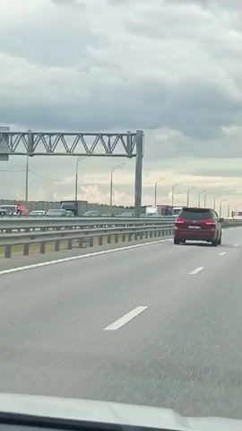 A dashcam view from a vehicle traveling on a multi-lane highway under an overcast sky. The highway has concrete barriers separating the lanes of traffic and guardrails on the left. Overhead structures with lights are visible. Multiple vehicles are on the highway, including cars and trucks. The car in front of the dashcam is a red SUV. Further ahead, a white sedan is visible, and to its left, other cars and trucks are in traffic. The road surface is asphalt with white lane markings. The environment appears to be an urban or suburban highway.