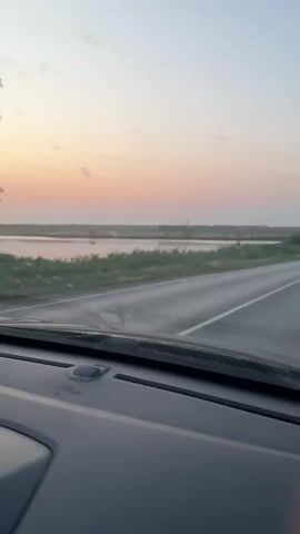 This is a first-person perspective video of someone driving on a paved road. The sky is a soft gradient of pink, orange, and pale blue, suggesting it is either sunrise or sunset. On the left side of the road, there is a body of water, likely a lake or marsh, bordered by green vegetation. On the right side of the road, there is more vegetation and a guardrail. The road itself is empty except for the vehicle the viewer is in. The dashboard of the vehicle is visible in the foreground, and the windshield is slightly dirty. The camera is pointed forward, capturing the road ahead and the surrounding landscape.