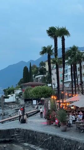 A vertical video shows a nighttime scene along a waterfront promenade. Several tall palm trees line the right side of the frame. To their right, a multi-story building with balconies and windows is visible, illuminated from within. Below the building and between the trees, an outdoor seating area for a restaurant is set up with tables and chairs, some occupied by people dining or socializing. Strings of lights hang above the seating area, and a Swiss flag is displayed in a potted plant. To the left of the palm trees and restaurant, a paved walkway runs along the water's edge. A single person stands near a speaker and a microphone setup on the left side of the walkway, seemingly performing or preparing to perform. A few other people are scattered along the walkway and on a wooden structure extending into the water on the far left. In the background, a body of water is visible, and beyond that, hills or mountains rise against a twilight sky. The overall atmosphere is one of a relaxed evening gathering.