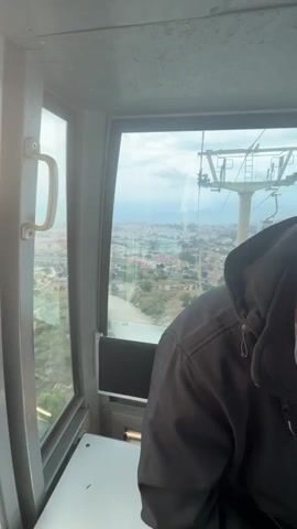 The video is shot from inside a cable car ascending over a city. The interior is simple, with a white seat and window frames. A young man, wearing a black jacket, a white t-shirt with colorful stripes, and black shorts, is seated. He looks at his phone and holds a small bottle of what appears to be beer or a similar beverage in his hand. A black GoPro camera on a small stand is positioned between his phone and the bottle. Outside the window, the city stretches out, with buildings and green spaces visible under a cloudy sky. A cable car tower is visible through the window as well.