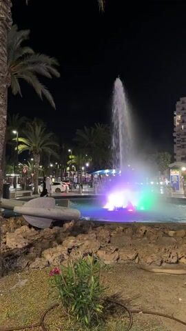 This vertical video captures a vibrant nighttime scene at a public fountain in Sant Antoni de Portmany, Spain. The fountain is the central subject, with a powerful jet of water shooting upwards, illuminated by a dazzling array of colorful lights in blues, purples, and greens. The water itself creates a luminous mist.

In the foreground, a sculpted object resembling a submarine or a torpedo lies partially submerged in the fountain's basin, nestled amongst rocky, natural-looking landscaping. A small green plant with pink flowers grows in front of this sculpture.

The setting is urban, with palm trees lining a street behind the fountain. Streetlights cast a warm glow on the scene, and the darkness of the night sky indicates it is nighttime. Several people are visible near the fountain, some appearing to be walking and observing the display. A vehicle, possibly a white car, is parked nearby, and in the background, there are buildings and hints of commercial activity, suggesting a lively area.

The atmosphere conveyed is one of lively, public enjoyment and spectacle. The colorful lighting of the fountain suggests a festive or tourist-oriented location, likely a town square or promenade. The overall impression is of a visually engaging and active urban space at night. No discernible text is visible in the frame.