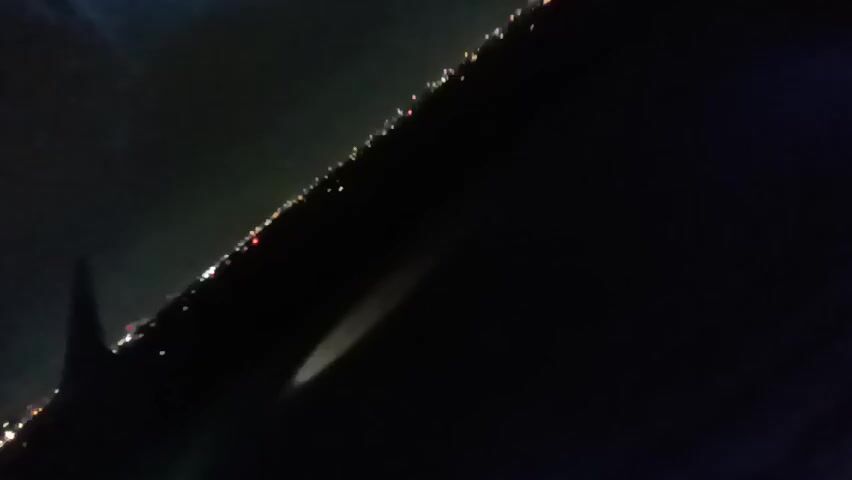 The video shows a dark scene, possibly filmed at night or in a low-light environment. A line of small, indistinct lights is visible along the upper left edge, possibly indicating a distant shoreline or cityscape. A blurry, light-colored shape, possibly a reflection, is visible in the lower center of the frame. A small red shape is visible on the left. The overall image is dark and lacks clear detail.