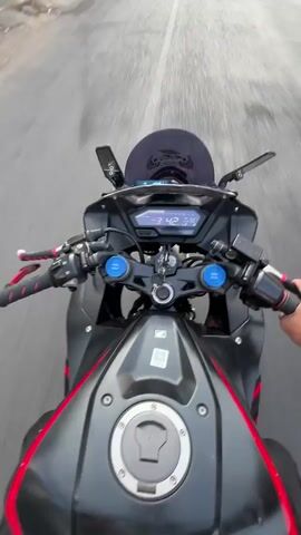 The video shows a first-person perspective of someone riding a motorcycle on a paved road. The motorcycle is black with red accents. The rider's hands are visible on the handlebars. The road is lined with buildings and trees, and other motorcycles and vehicles are present. The time of day appears to be daytime, possibly late afternoon due to the lighting. The motorcycle's dashboard is visible, displaying the speed. The rider is looking forward, and the scene is slightly blurred due to the motion of the motorcycle.