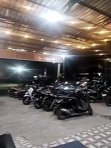 The video shows a parking area under a corrugated metal roof illuminated by strings of incandescent bulbs. Several motorcycles are parked in neat rows. A man in a red shirt stands near a small white structure in the background, appearing to be adjusting something. Outside the parking area, beyond a low wall, are trees and lights.