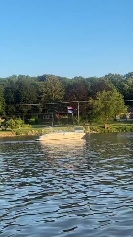 The video shows a white boat on a body of water, moving slowly towards the right side of the frame. The boat has a flag at the stern, which appears to be red, white, and blue. The water is choppy, with ripples and reflections. In the background, there is a green, tree-lined shore with several houses and a dock. The sky is clear and blue. The lighting suggests it is daytime, likely late afternoon or early evening due to the warm light. There are no people clearly visible on the boat or on the shore.