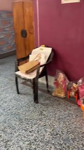 The video begins with a blurry shot of a wall with what appears to be religious figurines and an orange bucket. The camera then pans to a refrigerator and a large bag. The scene shifts to a room with a blue stool, a wooden cabinet, and a table with some items on it. The video continues to pan, showing a couch and a window with curtains. Finally, it shows clothes hanging on a clothesline in front of two arched windows. The overall scene depicts an interior living space, possibly a home, with various everyday objects and furnishings. There are no people visible in the video.