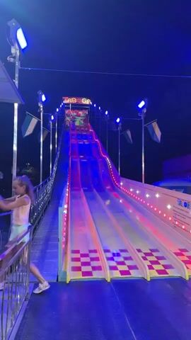 This media file captures a lively scene at an amusement park or fairground in Riccione, Italy, at night. The primary attraction is a large, multi-lane slide with bright, colorful lighting that shifts between red, pink, and green. The slide is elevated, with a steep incline leading down to a wide, flat landing area.

On the left side of the frame, a metal walkway with railings allows people to access the top of the slide. Two young girls are visible on this walkway, looking towards the slide with anticipation. One girl is wearing a light pink top and green shorts, while the other is in a white t-shirt and blue shorts.

A person, possibly a child, is seen sliding down the attraction, initially on a red section and then transitioning to the pink. Another person is walking up the ramp towards the slide, carrying a mat. The slide itself is yellow with a red and yellow checkered pattern on the lower sections, and it's illuminated by strings of lights.

Above the slide, a sign with colorful lettering is visible, though the text is partially obscured and difficult to read clearly. Flags in green, white, and blue are also mounted on poles around the attraction. The overall atmosphere is one of fun and excitement, typical of a nighttime fair with its vibrant lights and attractions. The time of day is clearly night, and the weather appears to be clear, suitable for outdoor amusement.