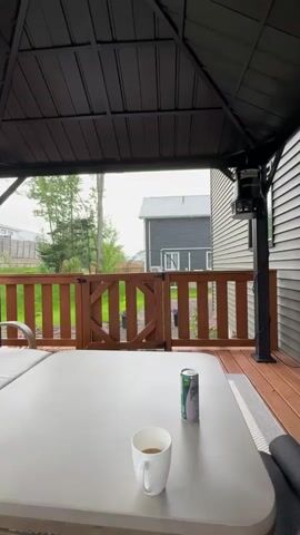A video shows a person sitting at a table on a covered deck as it begins to rain. In the foreground, a white mug with a beverage and a can of sparkling water sit on a light-colored table. A wooden fence with a gate stands behind the table, and beyond the fence is a yard with grass, trees, and a dark-colored house. The rain intensifies throughout the video, creating visible streaks in the air.