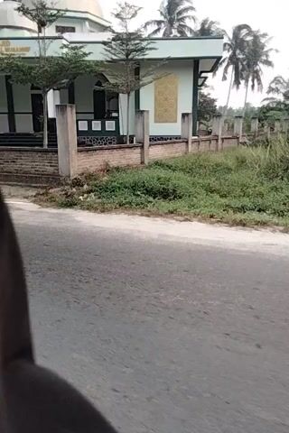 The video shows a close-up, low-angle view of a paved road, likely in a rural or suburban area. To the left of the road, there is a building with a distinctive architectural style, featuring arched doorways and decorative elements, suggesting it might be a mosque or similar religious structure. A fence runs along the property line, and vegetation, including trees and grass, is visible. The road surface appears somewhat worn, with some areas of discoloration. The camera is moving, indicating that the footage was captured from a moving vehicle, possibly a motorcycle, given the partial view of a wheel and the dynamic perspective. The overall environment appears to be an outdoor setting, with natural light suggesting daytime. The scene is devoid of any clear human subjects actively interacting within the frame, although parked motorcycles are visible near the building.