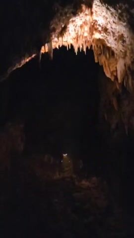 The video shows the interior of a dark cave with illuminated stalactites hanging from the ceiling. The camera pans downward, revealing the rocky floor and walls of the cave. There are small, dim lights visible in the distance, possibly indicating a path or other explorers. The overall impression is one of a natural underground environment.