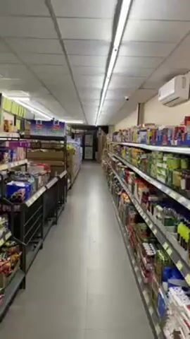 The video shows a walk-through of an aisle in a grocery store or supermarket. The aisle is long and narrow, with shelves on both sides filled with various food and household products. The shelves on the left side are metal and display items such as snacks, cookies, and boxed goods. The shelves on the right side appear to be stocked with canned goods, sauces, and condiments. The lighting in the store is bright and fluorescent. The floor is gray and clean. There are no people visible in the video. The camera moves slowly forward through the aisle.