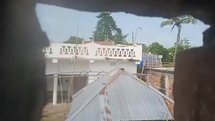 The video shows a view through a rough opening in a brick wall. In the background, there is a white building with a decorative railing on its upper level. To the right of this building, there is a palm tree. In front of the white building, there is a corrugated metal roof. The sky is overcast.