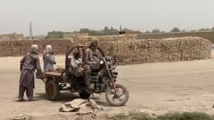 The video shows a motorized three-wheeled vehicle, reminiscent of a motorcycle with a flatbed trailer attached, parked on a dirt road in what appears to be a brickyard. Seated on the vehicle are two individuals; the driver is wearing a turban and sunglasses. Another person is seated in the trailer section. Two other people are standing nearby.  In the background are stacks of bricks, a tall chimney, and some sparse vegetation. The overall environment is dry and dusty.