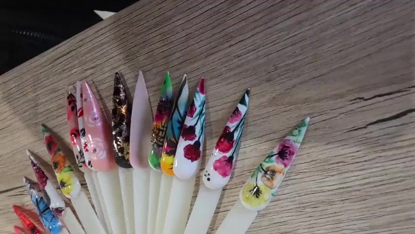 A person's hand with a fingernail is visible in the upper right corner of the frame, hovering over a collection of artificial nails. The artificial nails are displayed on white sticks, fanned out on a wooden surface. Each nail is intricately designed with various patterns and colors, including floral designs, abstract patterns, glitter, and solid colors. The lighting is bright and focused on the nails, casting subtle shadows on the wooden surface.