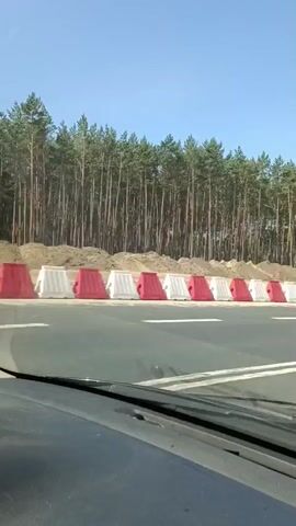 The video captures a construction scene near Mała Nieszawka, Poland, on a clear, sunny day. The perspective is from inside a vehicle, looking out at a highway under construction. Red and white plastic barriers line the side of the road, demarcating the construction zone. Beyond the barriers, a dense forest of pine trees forms a backdrop, with a sandy embankment.

The main activity is earthmoving. A large yellow Caterpillar articulated dump truck is positioned on the embankment, likely waiting to be filled. An excavator, also yellow, is actively digging into the sand and appears to be loading the dump truck. The excavator's boom is extended, and its bucket is visible, filled with dirt. The scene suggests ongoing road work or land preparation.

A prominent yellow, oval-shaped object is visible in the sky in the early frames, which may be a balloon or other airborne object. Traffic cones with red and white stripes are scattered along the edge of the active construction area. The road itself is paved with white lane markings. There is no visible text within the video. The overall impression is one of active, large-scale construction in a rural, wooded environment under bright daylight.