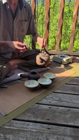 A person sits cross-legged on a wooden deck, preparing tea. They wear a checkered shirt and brown pants. In their left hand, they hold a small, round, brown object, possibly tea leaves or a tea cake. With their right hand, they pour liquid from a dark brown teapot into a smaller, similar-colored teapot. In front of the teapots, arranged on a mat, are three light-colored ceramic bowls with speckled rims. Behind the teapots, a portable gas stove is visible. The background shows a lush, green forest with tall trees and a wooden railing. The lighting suggests it is daytime, possibly late afternoon due to the shadows.