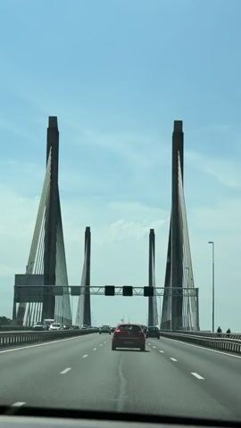 This is a first-person perspective video taken from inside a vehicle as it drives on a highway towards a large cable-stayed bridge. The sky is blue with some clouds. The bridge has four prominent towers and many cables supporting the roadway. There are multiple cars on the highway, with one red car directly in front of the camera vehicle. There are traffic signs visible above the highway. The overall impression is one of driving on a sunny day towards a significant piece of infrastructure.