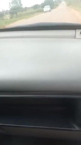 The video captures a first-person perspective from inside a vehicle, looking out through the windshield at a rural road. The dashboard of the car is prominently in the foreground, a light gray plastic surface. Beyond the dashboard, a paved road stretches into the distance, curving slightly to the right. Trees and green vegetation line the sides of the road, and in the far distance, a white car is visible traveling away from the viewer. The sky is overcast with clouds. The car appears to be in motion, as indicated by the slight blur of the distant scenery. The overall impression is of a drive through a natural, possibly undeveloped, landscape.