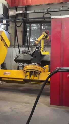 A robotic arm on a yellow industrial fixture is moving a dark metal component. The robot is yellow and has multiple joints. The fixture it is attached to is also yellow and has the "VIRS" logo on it. The background is an industrial setting with red curtains, windows, and other machinery. A black cable is visible in the foreground, extending into the frame from the right. The component the robot is holding is black and appears to be metallic. The lighting is artificial and bright.