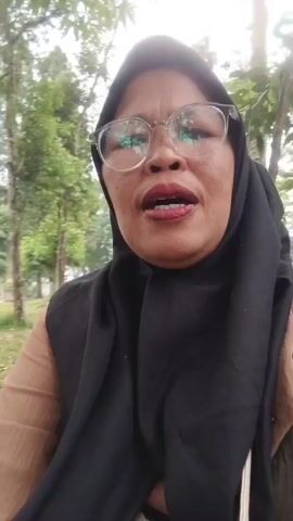 The video opens with a close-up of a woman wearing a hijab and glasses, speaking directly to the camera. The background shows a park-like setting with trees and a slightly overcast sky. As the camera pans and zooms out, it reveals a grassy area with several tents set up, indicating a camping or picnic event. Multiple groups of people are visible in the background, some sitting on the ground, others interacting with each other. One group appears to be having a picnic, with blankets spread out and food items visible. Another group is gathered near a tent. The overall atmosphere suggests a casual outdoor gathering. The focus then shifts to the environment, showcasing the trees, fallen leaves on the ground, and the general layout of the park.