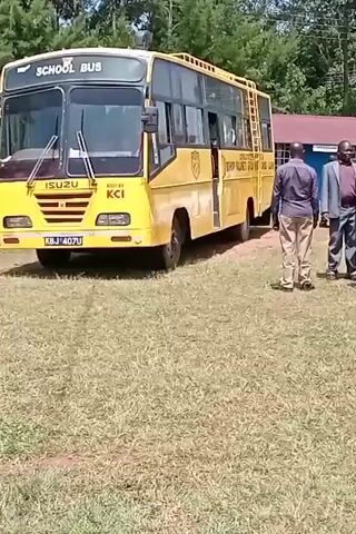Here is a concise description of the scene from the video:

The video shows a yellow school bus parked on a grassy area next to a group of men. The bus has "SCHOOL BUS" and "ISUZU" printed on it, as well as what appears to be the name of a school. The men are standing and talking near the bus and other vehicles are also visible in the background. The background includes trees, a building with a red roof, and other vehicles.  The day appears sunny.