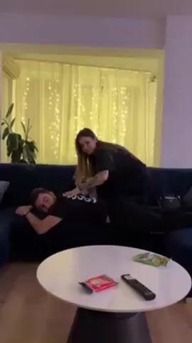 A woman is sitting on a man's back on a dark blue couch, giving him a massage. The man is lying prone on the couch, wearing a black t-shirt with white writing and black pants. The woman is wearing a black t-shirt and black pants. There is a round, white coffee table in front of the couch. On the table, there is a remote control, a packet of crisps, and a red packet. Behind the couch, there is a large window covered by a sheer white curtain with yellow fairy lights hanging behind it. To the left of the couch, there is a green plant. The room appears to be a living room.