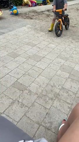 A child on a balance bike rolls forward on a paved surface. The child is wearing a gray t-shirt, yellow boots, and is positioned in the center of the frame. The balance bike is black and orange. The paved surface is made of square concrete tiles. In the background, there is a sandy area with various children's toys, including a yellow and blue toy, a pink toy, and a green and yellow toy. The perspective is from a seated position, with a person's legs visible in the foreground.