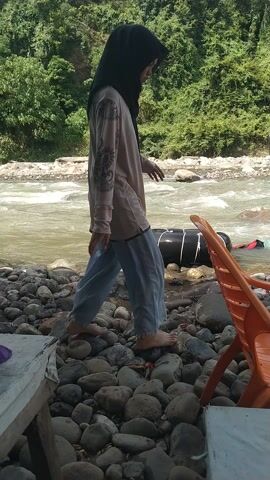 This is a video of people on a river in Meurah, Indonesia. The scene is a rocky riverbank with lush green trees on one side. The river appears to be flowing rapidly, with some white water visible.

There are at least three people visible in the water. One person, wearing a dark hijab and dark clothing, is standing in the river near a large, black inflatable raft. They appear to be interacting with the raft. Another person, wearing a light-colored shirt and light pants, is partially visible standing further up the river, also appearing to be in the water. A third person, wearing a light blue shirt, is visible further upstream, also in the river. The overall mood seems relaxed and possibly recreational.

An orange plastic chair is placed on the rocky bank, suggesting a place where people might sit and watch or rest. The time of day appears to be daytime, with bright sunlight casting shadows, indicating fair weather. The presence of the river and the surrounding vegetation points to a natural, possibly rural or semi-rural, outdoor setting. There is no visible text in the video.