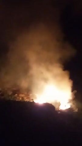 The video shows a fire burning in the dark, with orange flames and smoke rising upwards.  The camera is shaky, making it difficult to discern the exact nature of what is burning, but it appears to be an outdoor fire. The end of the video becomes a blurred, close-up view of the flames and smoke.