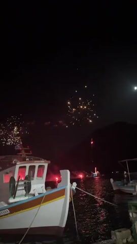 The video captures a nighttime scene at a harbor with fireworks illuminating the dark sky. A white fishing boat with a blue and orange stripe along its hull is docked in the foreground, tethered by ropes to a stone quay. The boat has a mast with a Greek flag and a red light visible on its deck. The water reflects the vibrant colors of the fireworks, which are bursting in red, white, and yellow. In the background, other boats are moored, and distant lights suggest a settlement along the water's edge. The overall atmosphere is festive and celebratory.