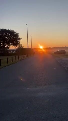 A paved road stretches towards a sunrise in the distance. A lone car is parked to the right of the road. A tree stands on the left side of the road, with a wooden fence bordering the road. The sky is clear with a slight orange hue from the rising sun. The sun is low on the horizon, casting a bright glow across the landscape.