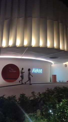 The video shows a nighttime view of a modern building, likely a hotel or resort, with illuminated signage for "SEEN." The building features a curved entrance with recessed lighting and the name "AVANI" displayed. A couple is walking in front of the building, and there are plants in the foreground. The camera pans to the right, revealing more illuminated "SEEN" signs, with one advertising "SEEN Beach Club" and another advertising "SEEN Eatery." The last sign features images of food and people.  A building with a red, triangular pattern is visible in the background.