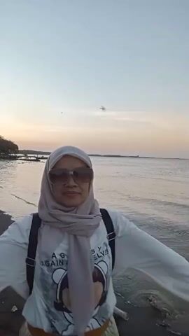 A full-shot video shows a woman standing on a dark, sandy beach with her arms outstretched, facing the ocean. The sun is setting, casting a warm glow on the horizon. The woman is wearing a hijab, glasses, a white long-sleeved shirt with a graphic print, and a long skirt. She is carrying a backpack. The beach is littered with trash. In the background, there are a few small buildings and trees along the coastline, and a body of water. The camera pans slightly to the right and left, following the woman's movement.