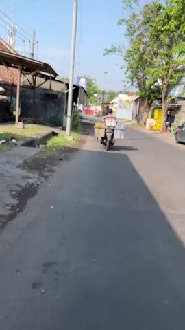 A motorcycle with a large cargo box is seen driving down a paved road. The motorcycle is a dark color and the cargo box appears to be red and white. The road is lined with buildings on both sides and trees are visible in the background. The sky is blue and the sun is shining. A person is riding the motorcycle. There are no other people visible in the immediate vicinity.