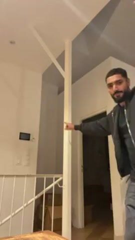 The video shows a man standing in a room with a staircase and a doorway leading to a dark room. He is wearing a black hooded jacket, a black shirt, and faded grey jeans. He has a short beard and dark hair. He starts by pointing towards the right, then spreads his arms out to the sides and dances. A second person, who is Black, wearing sunglasses and a cream-colored hoodie with a graphic on it, joins him, putting their arms around him. The room is well-lit, with white walls and a modern design. There is a light switch on the wall and cardboard boxes are visible in the dark room through the doorway.
