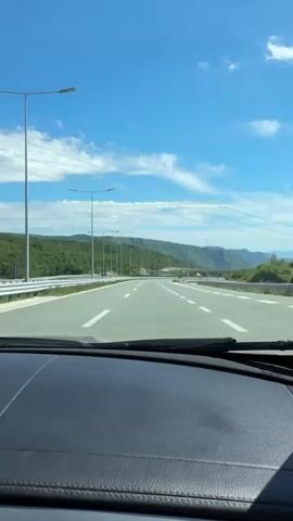 A first-person view from inside a car shows a highway stretching into the distance. The sky is bright blue with scattered white clouds. On either side of the highway are green, rolling hills. The road is paved and has white lane markings. Streetlights line the left side of the road. There are power lines overhead. The car's dashboard is visible in the foreground.