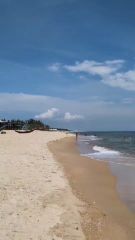 This video depicts a tropical beach with light blue skies and scattered white clouds. The foreground is dominated by a wide expanse of pale sand. The ocean, with gentle waves lapping at the shore, fills the right side of the frame. Several people are visible in the water, some appearing to be swimming or wading. Along the left side of the beach, closer to the tree line, a small, dark-colored boat is pulled ashore, and a few structures that resemble buildings or shelters are present. The overall scene conveys a sense of a serene and sunny day at the beach.